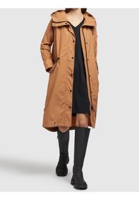 Parka impermeabile marrone con cappuccio, bottoni a pressione e tasche laterali. Indossata sopra un vestito nero con stivali di gomma neri alti fino al ginocchio.