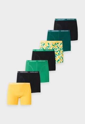 Slanke donkerhuidige man staat blootsvoets, draagt gele boxershorts met groene blad- en citroenpatronen tegen een effen witte achtergrond.
