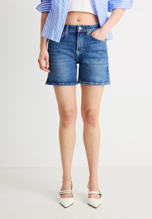 Frau trägt blaue Jeansshorts, weiße spitze Heels mit doppelten Riemen, ein kurzes weißes Top und ein blau gestreiftes Hemd zum Knöpfen.
