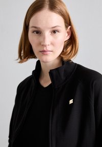Crna jakna na zip s visokim ovratnikom, koja ima mali zlatni emblema. Nosena preko crne majice s V izrezom. Glatka tekstura.