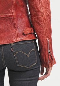 Red leather jacket with a perforated texture, adjustable strap detail, and zippered cuff, paired with dark denim jeans featuring a distinctive back pocket stitch.