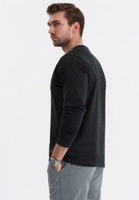 Schwarzes Langarmshirt aus weichem Stoff, mit rundem Ausschnitt und enganliegenden Ärmeln. Kombiniert mit grauen Nadelstreifenhosen.