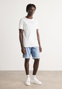Fehér pamut póló, rajta a "Calvin Klein Jeans" felirattal, világoskék farmer rövidnadrággal párosítva, a szárak felgyűrve, és fehér sportcipő világos zoknival.