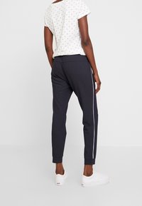 Person klädd i mörka slim fit-byxor med vita sidostråk, vit tryckt kortärmad skjorta och vita sneakers, stående mot en neutral bakgrund.