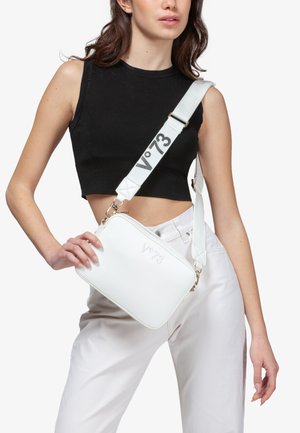 Donna che indossa un top corto nero senza maniche e pantaloni bianchi, che tiene una borsa a tracolla bianca con la scritta "V° 73" sulla tracolla e sul davanti.
