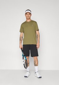 Camiseta atlética verde oliva, calções pretos, faixa branca na cabeça, segurando uma raquete de ténis. Apresenta tatuagens nos braços, meias brancas e ténis.