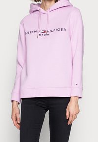 Helle rosa Kapuzenjacke mit Kordelzugkapuze, mit schwarzem Text "TOMMY HILFIGER EST. 1985" und Logo, aus weichem Stoff, lockere Passform.