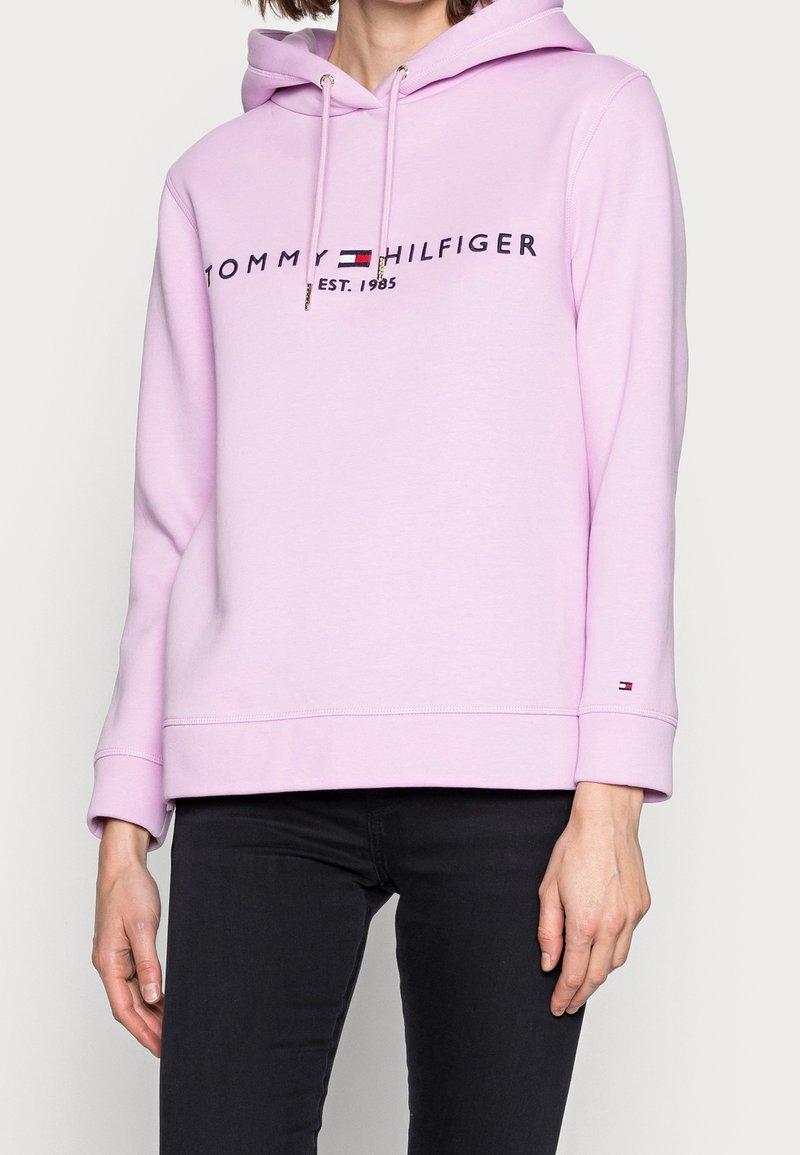 Helle rosa Kapuzenjacke mit Kordelzugkapuze, mit schwarzem Text "TOMMY HILFIGER EST. 1985" und Logo, aus weichem Stoff, lockere Passform.