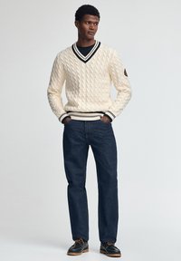 Kremowy sweter z warkoczowym splotem, z dekoltem w serek i czarnym wykończeniem, w zestawie z ciemnoniebieskimi dżinsami i ciemnymi butami. Szczegóły w postaci ściągaczy na mankietach i u dołu.