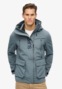 Grå vindjakke med lynlås foran, hætte, to sidelommer, to brystlommer og et markant "SUPER" logo på fronten.