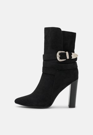 Czarny zamszowy botek na wysokim obcasie z zaostrzonym noskiem, ozdobną srebrną klamrą i paskiem wokół cholewki.