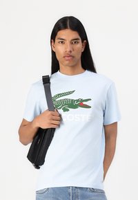 Šviesiai mėlyna medvilninė marškinėlių su didele žalia krokodilo logo ir balta "LACOSTE" užrašu. Modelis laiko mažą juodą krepšį su petnešėlėmis.