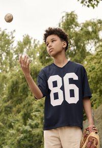 Granatowy sweter z dużą białą cyfrą "86" z przodu, noszony przez dziecko trzymające rękawicę baseballową. W tle widać zieleń i pochmurne niebo.