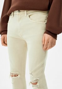 Persona con jeans rotos beige y una camiseta de manga larga marrón, con las manos relajadas a los lados, cortada a mitad del muslo.