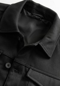 Sort denimjakke med klassisk krave, knaplukning og en forlommer. Teksturen virker robust med en glat inderforing.