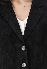 Blazer noir texturé avec des motifs floraux, design croisé, et trois boutons floraux en argent, porté sur un haut blanc à côtes.