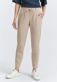 Pantalones beige ajustados con cintura elástica y cordón, que cuentan con bolsillos laterales y una textura de tela mate y suave. Llevados con zapatillas blancas.