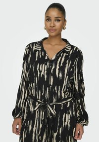 Chemise boutonnée noire et crème à rayures avec des manches longues et une taille nouée. Tissu doux au toucher lisse et col classique.