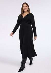 Černé šaty s dlouhým rukávem a uzlovým detailem v pase. Látka má žebrovanou texturu a délku do poloviny lýtek, doplněné černými botami.
