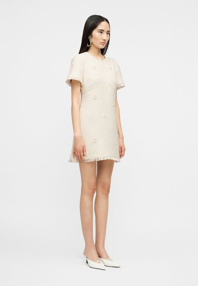 Νεαρή γυναίκα όρθια, φοράει ένα κρεμ φόρεμα με κοντά μανίκια και υφή, διακοσμημένο με μικρά λουλουδάτα σχέδια, με φουντωτές άκρες, μεγάλα χρυσά κρίκους σκουλαρίκια και λευκά τακούνια.