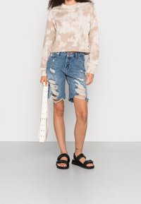 Person som har på sig en beige tie-dye långärmad skjorta, slitna knälånga denimshorts, svarta sandaler och håller en vit bälte eller rem.