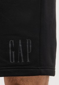 Μαύροι υφασμάτινοι σορτς με τη λέξη «GAP» τυπωμένη κατακόρυφα κοντά στο τελείωμα στο πόδι ενός ατόμου.