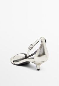 Srebrne metaliczne sandały na wysokim obcasie z paskiem i klamrą, odkrytymi palcami, zakrzywionym wzorem i cienkim obcasem. Gładka faktura na powierzchni.