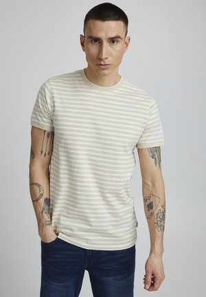 Stripet t-skjorte i krem og beige, korte ermer, rund hals, laget av mykt stoff. Har synlige arm tatoveringer og jeans.