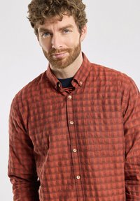 Chemise à carreaux rouges à boutons, en tissu texturé, avec un col et une poche poitrine unique, ornée de boutons de couleur claire et de motifs subtils.