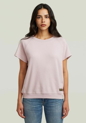 Giovane donna con lunghi capelli scuri che indossa una maglietta a maniche corte rosa chiaro e jeans blu su uno sfondo chiaro e uniforme.
