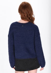 Donkerblauwe fluffy trui met een ronde hals, verlaagde schouders en brede mouwen. De trui heeft een korte zoom en een kleine metalen accent aan de achterkant.