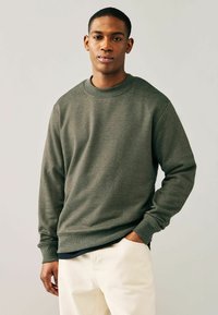 Olivgrön sweatshirt med rundad halsringning, långa ärmar, ribbade muddar och midja. Buren över en mörk tröja och parat med ljusa byxor.