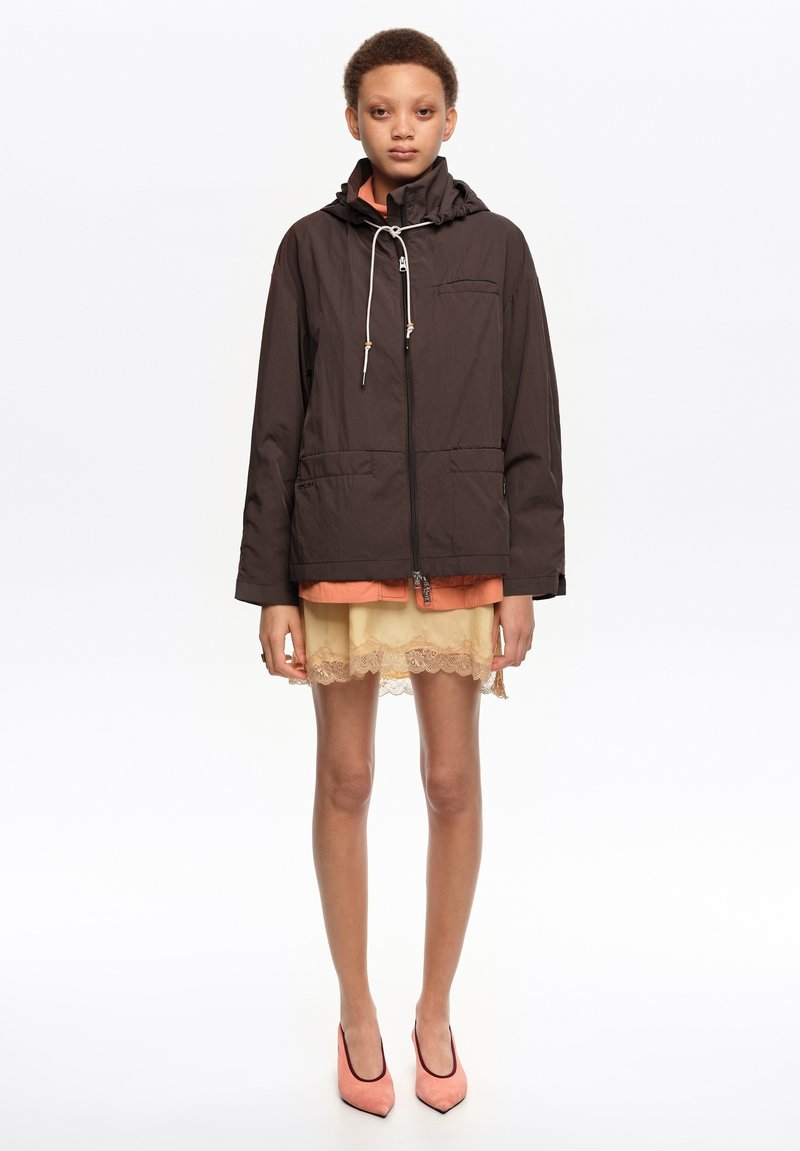 Isik, kes kannab musta kapuutsiga jope oranži topi ja kreemika pitsäärega seeliku peal, seljas korallpunased teravate ninadega kingad, seisab valge tausta ees.