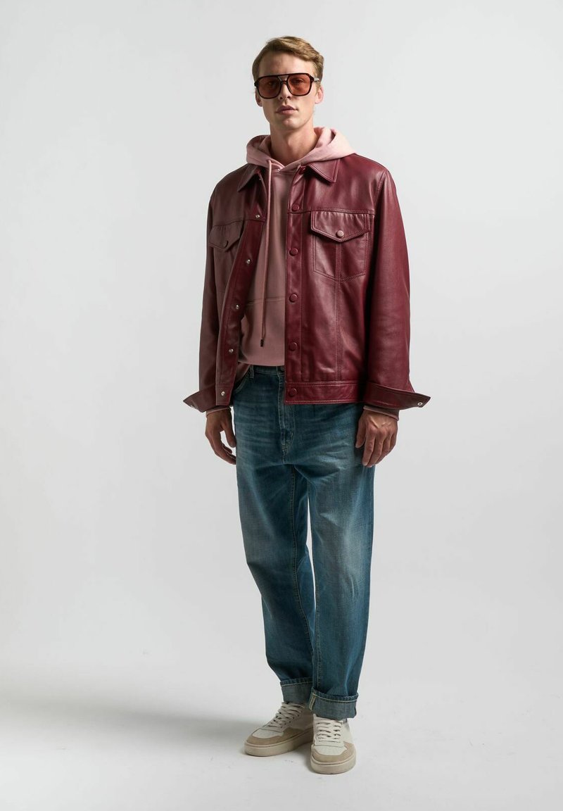 Giubbotto in pelle bordeaux sopra un felpa leggera rosa, abbinato a jeans azzurri e sneakers beige. Occhiali da sole marroni oversize completano il look.