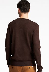 Mann mit kurzen dunkelbraunen Haaren, der einen schlichten dunkelbraunen Pullover mit langen Ärmeln und hellbraune Hosen trägt und mit dem Rücken zum Betrachter vor weißem Hintergrund steht.