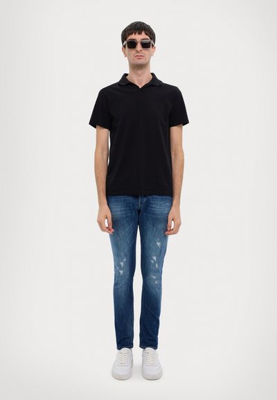 Musta polo-paita, siniset kuluneet slim-fit farkut ja valkoiset tennarit. Malli, jolla on ylikokoiset mustat aurinkolasit, seisoo yksinkertaista taustaa vasten.