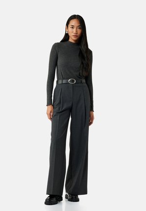 Vrouw met lang zwart haar, gekleed in een donkergrijze aansluitende top met lange mouwen, grijze wijde broek met hoge taille en een zwarte riem, en zwarte schoenen.