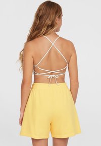 Pantaloni shorts gialli in cotone con vita elasticizzata e cordino, abbinati a un top chiaro con schiena scoperta e spalline incrociate.