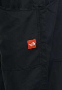 Μαύρο ύφασμα με ορατά ραφές και μια μικρή κόκκινη ορθογώνια ετικέτα που εμφανίζει το λογότυπο "THE NORTH FACE" με λευκά γράμματα.