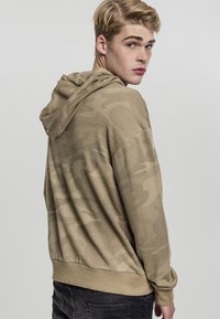 Beige hettegenser med et subtilt camouflasjemønster, myk tekstur, avslappet passform, ribbestrikkede ermer og linning. Bakview vist.