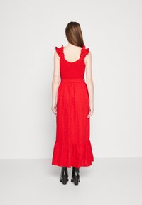 Punainen mekko, jossa on röyhelöillä varustetut olkaimet, rypistetty yläosa ja kerroksellinen hame. Valmistettu teksturoidsesta kankaasta, joka sisältää muotoillun vyötärön ja nilkkapituuden.