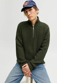 Pull en tricot vert foncé avec un col zippé mi-haut, présentant des motifs texturés et des poignets côtelés. Porté sur un jean bleu clair.