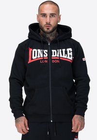 Sudadera con cremallera negra hecha de tela suave, con una capucha amplia, bolsillos frontales y un audaz logo en blanco y rojo de "Lonsdale" en el pecho.