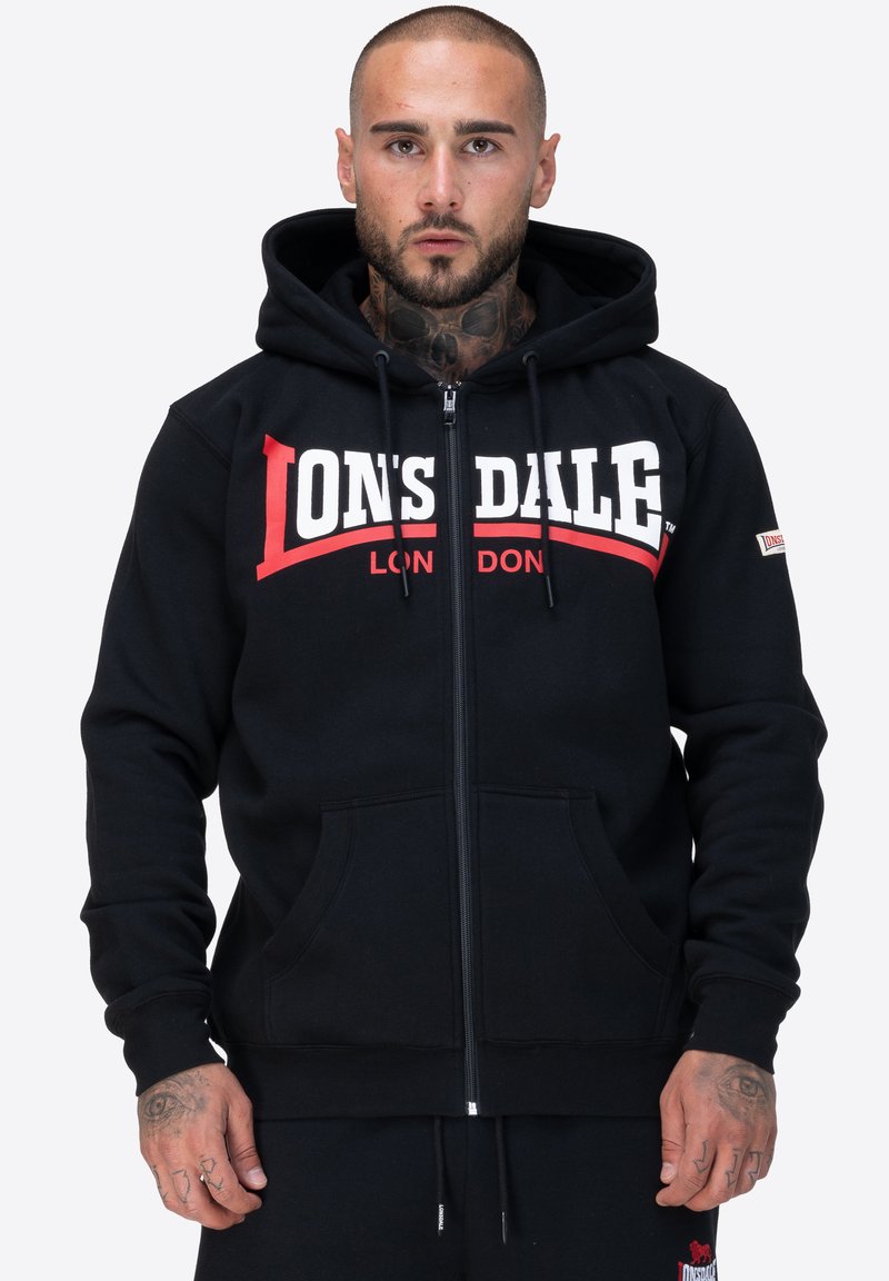 Sudadera con cremallera negra hecha de tela suave, con una capucha amplia, bolsillos frontales y un audaz logo en blanco y rojo de "Lonsdale" en el pecho.