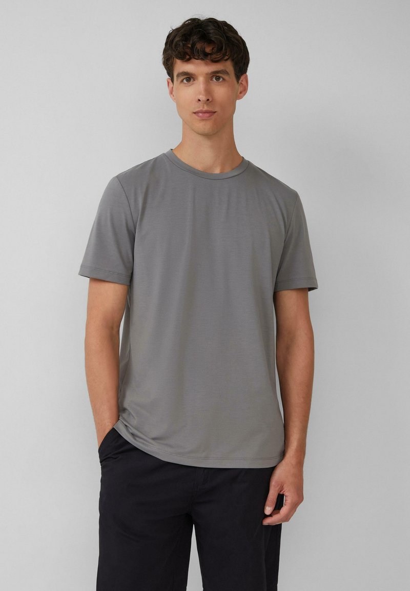 Grijze kraagloze t-shirt met korte mouwen, zachte textuur en slim fit. Model draagt donkere broeken, handen in de zakken tegen een lichte achtergrond.