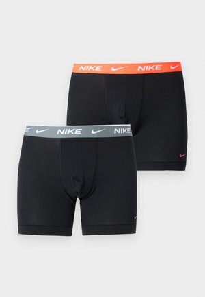 Homme torse nu debout pieds nus sur un tapis portant un boxer Nike noir avec une ceinture orange, regardant vers sa gauche sur un fond uni.