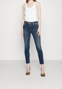 Mujer con top blanco, jeans ajustados azules con cinturón de hebilla dorada y tacones de serpiente de pie contra un fondo liso.