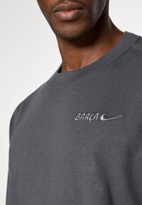 Grå bomullssvetter med rund hals, brodert "BARÇA" og Nike-logo i hvitt på brystet. Myk tekstur.