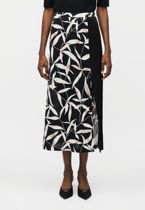 Γυναίκα που φοράει μαύρη midi φούστα με λευκά και μπεζ σχέδια φύλλων και πλαϊνή σκίσιμο, σε συνδυασμό με μαύρα ψηλοτάκουνα παπούτσια με μυτερή μύτη.