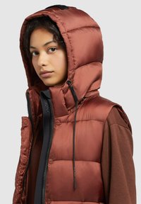 Padded Weste in Bronze mit hohem Kragen und Kapuze. Sie verfügt über einen Reißverschluss vorne, Zugbänder und eine Mischung aus Texturen. Sichtbare Details in schwarz.